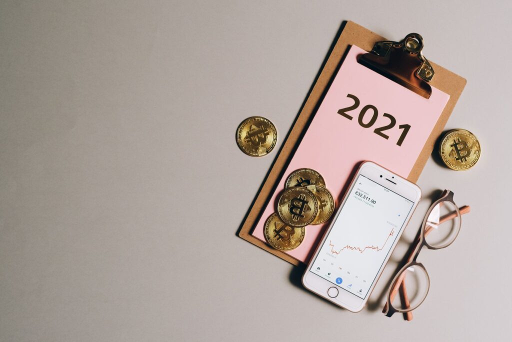 Flat lay of bitcoins, smartphone, eyeglasses, and clipboard with 2021 text, symbolizing cryptocurrency trends.