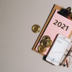 Flat lay of bitcoins, smartphone, eyeglasses, and clipboard with 2021 text, symbolizing cryptocurrency trends.