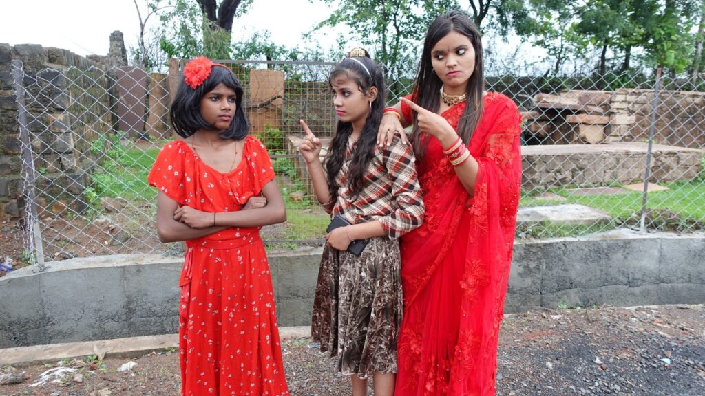 Three young women in vibrant dresses show expression against a fence backdrop.