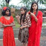 Three young women in vibrant dresses show expression against a fence backdrop.