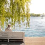Serene scene of an elderly couple sitting on a bench by a lakeside under a willow tree.