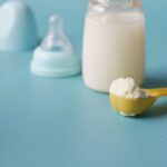 Close-up of baby milk formula powder in a yellow scoop with a bottle on a blue background.