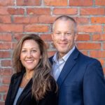 A cheerful middle-aged man and woman in business attire posing against a brick wall, depicting professionalism and partnership.