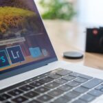 Close-up of a laptop displaying Adobe software icons, beside a camera on a desk.