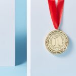 Close-up of a gold medal with a red ribbon hanging against a light blue background.