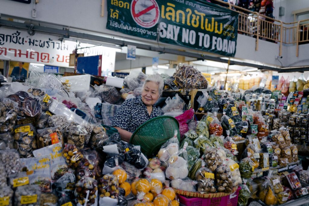 Elderly woman in an indoor market stall selling a variety of fresh produce and goods.