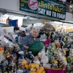 Elderly woman in an indoor market stall selling a variety of fresh produce and goods.