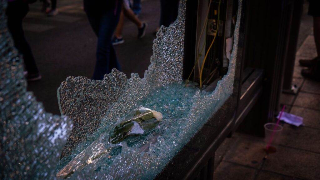 A shattered glass window on an urban street with a rose inside, capturing a moment of chaos and tranquility.
