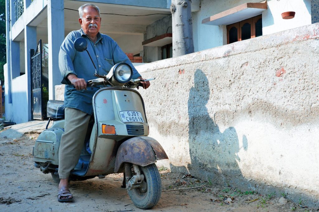 Elderly man with mustache riding a vintage scooter outdoors on a sunny day.