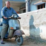Elderly man with mustache riding a vintage scooter outdoors on a sunny day.