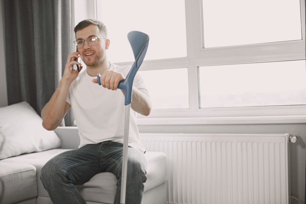 Adult man sitting on couch using crutch, talking on phone in a well-lit room.