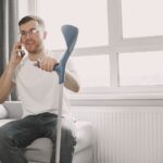 Adult man sitting on couch using crutch, talking on phone in a well-lit room.