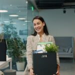 Happy businesswoman carrying belongings and plant into a modern office.
