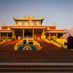 Golden sunlight illuminates a Buddhist temple in Sidhbari, with a monk standing nearby, creating a serene atmosphere.