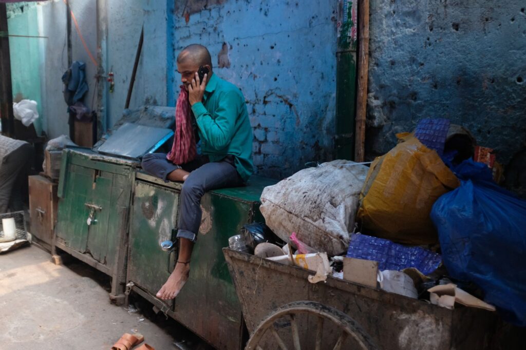 Man sitting in an urban slum using a smartphone amidst garbage and debris, New Delhi, India.