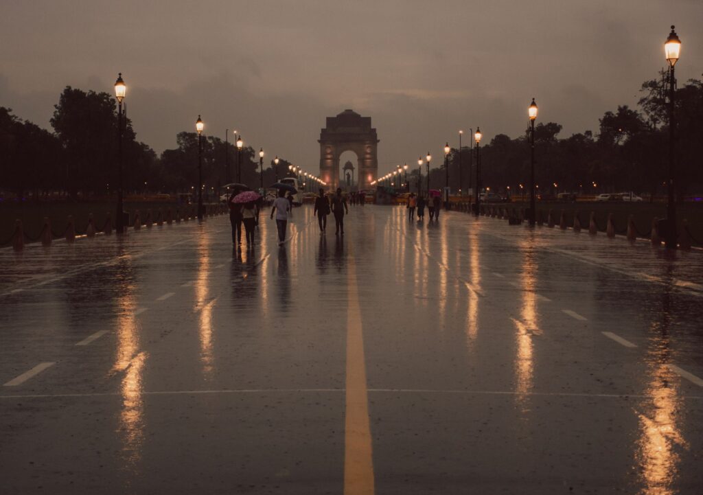 Moody evening scene at India Gate with reflections on the wet road and people walking.