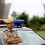 Indian police officer with arms crossed standing by a patrol car outdoors, showcasing authority and law enforcement presence.