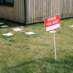A red 'House for Rent' sign stands on a grassy lawn beside a wooden house exterior.