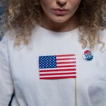 A woman with curly hair holding an American flag and wearing a vote pin against a blue background.