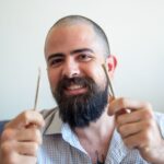 A cheerful bearded man with a checkered shirt holding two cannabis joints indoors.