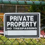 Close-up of a private property no trespassing sign on a chain-link fence with greenery in the background.