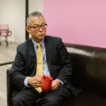 An Asian businessman in a suit, sitting on a sofa with a red mug in a modern office setting.