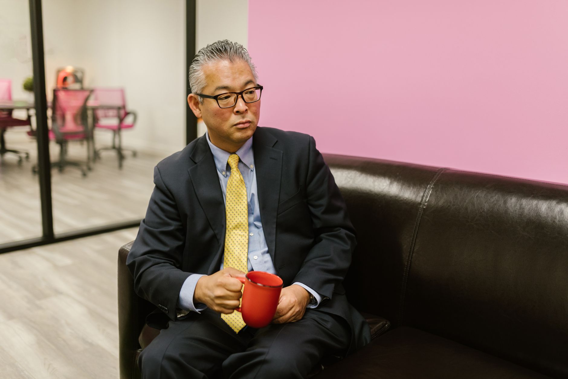 An Asian businessman in a suit, sitting on a sofa with a red mug in a modern office setting.