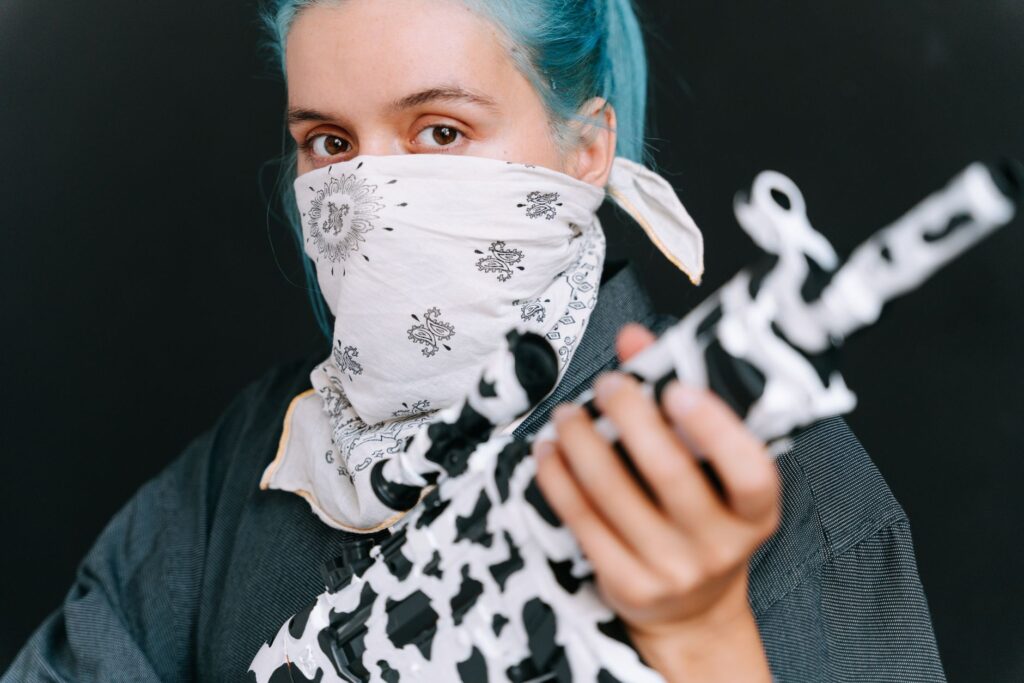 A person with blue hair and a bandana holding a black and white rifle, conveying protest themes.