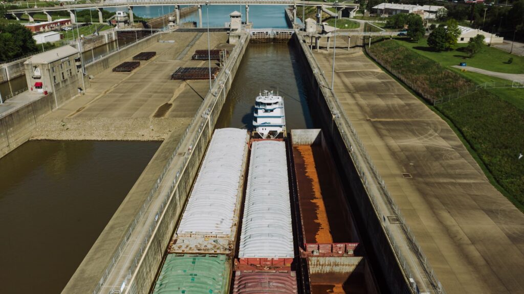 From above of old industrial transport carrying heavy containers with rusty surface on water channel near green meadow in harbor