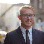 Smiling bearded male employee wearing formal clothes and eyeglasses looking away while standing on street against city downtown