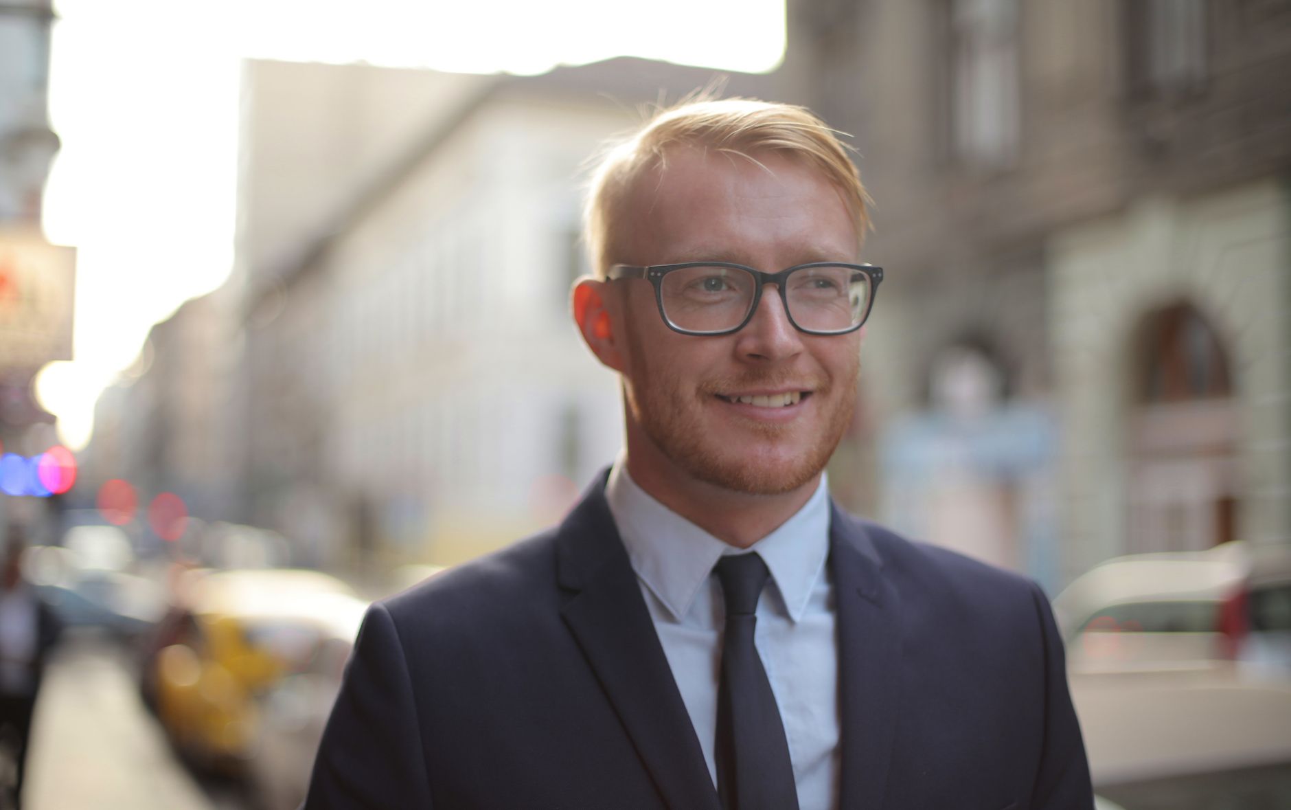 Smiling bearded male employee wearing formal clothes and eyeglasses looking away while standing on street against city downtown