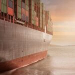 A colossal cargo ship loaded with containers navigates through calm waters against a vibrant sunset sky.