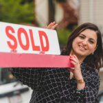 Smiling realtor holding a 'Sold' sign in front of a house. Perfect for real estate marketing.