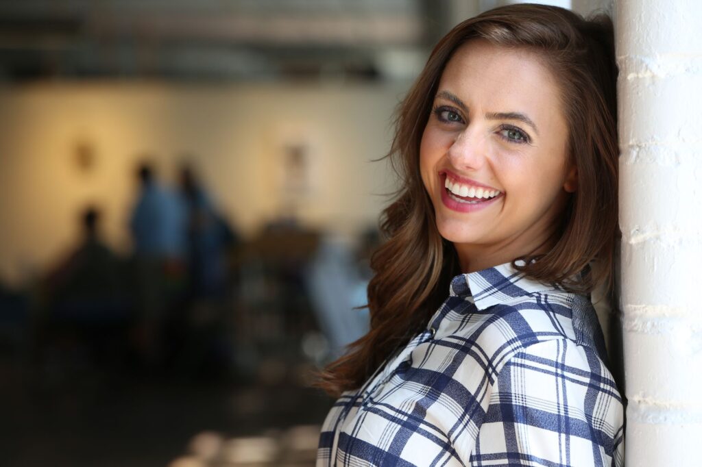 Portrait of a smiling woman in a plaid shirt leaning against a wall indoors.
