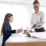From below of smiling female in formal wear and glasses tutor explaining homework task to little girl in creative place