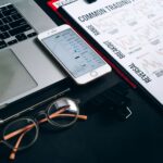 Workstation with laptop, smartphone, eyeglasses, financial charts, and pen for trading analysis.