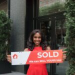 Confident real estate agent holds 'SOLD' sign in front of building, symbolizing success.