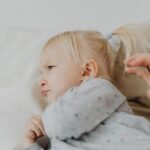 Cute blonde child with ponytail sulking on a soft bed indoors.