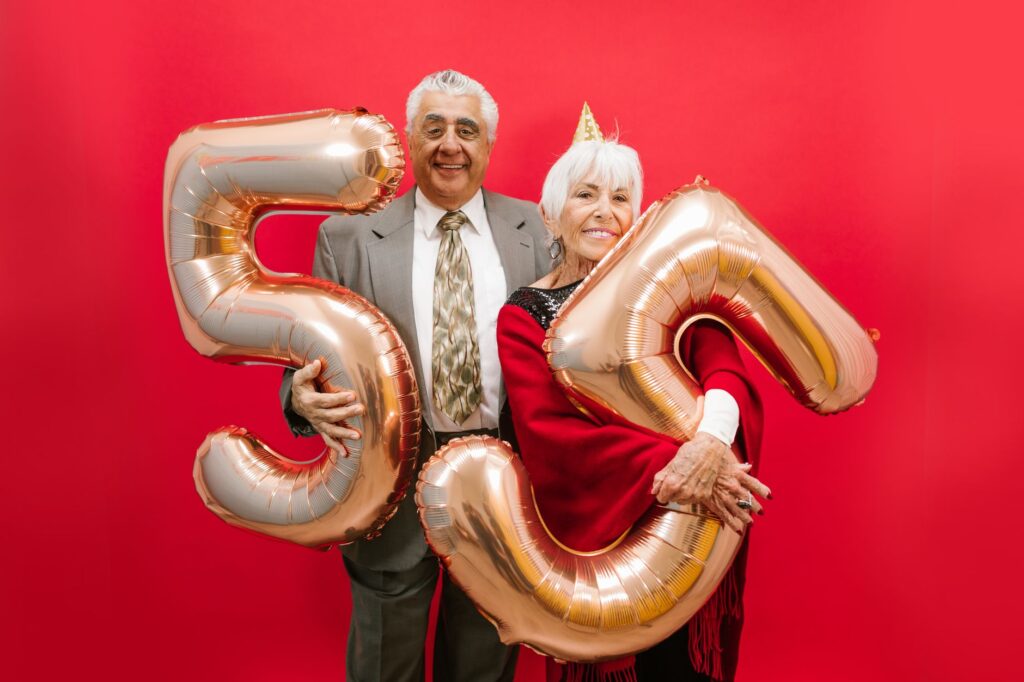 Happy senior couple in formal attire celebrating with golden balloons against a bright red backdrop.