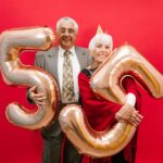 Happy senior couple in formal attire celebrating with golden balloons against a bright red backdrop.