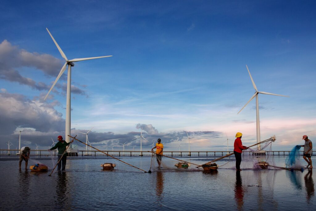 A group of fishermen cast nets near wind turbines set against a vibrant sunset sky, showcasing sustainable energy.
