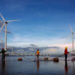 A group of fishermen cast nets near wind turbines set against a vibrant sunset sky, showcasing sustainable energy.