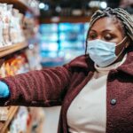 African American female buyer in protective mask picking food from shelf in grocery store