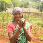 Elderly woman farmer smiling in a vibrant countryside garden with green crops.