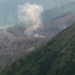 A dramatic view of a volcano crater emitting smoke amidst verdant mountains.