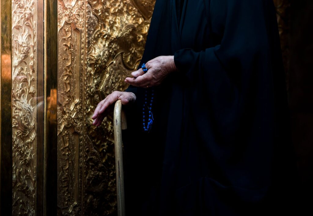 An elderly person holds a rosary and walking stick against a golden ornate wall in Karbala, Iraq.
