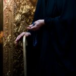 An elderly person holds a rosary and walking stick against a golden ornate wall in Karbala, Iraq.