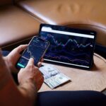 A person using a smartphone and tablet for stock market analysis, with graphs and cash visible.