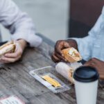Two friends enjoying wraps and sandwiches at a rustic outdoor wooden table, sharing a casual lunch.