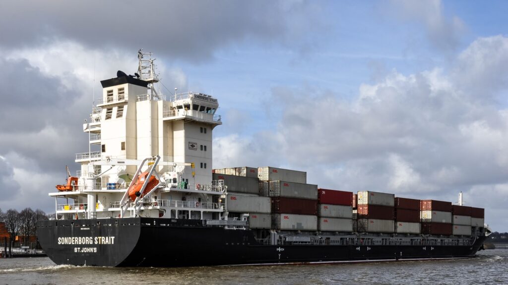 Container ship navigating Hamburg harbor with stacked cargo under a cloudy sky.